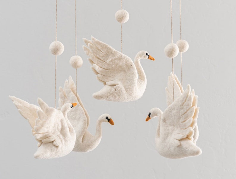 Puede incluir: Un m&oacute;vil de fieltro blanco con cuatro cisnes de fieltro blanco y cinco bolas de fieltro blanco colgando de un techo blanco.