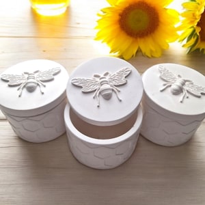 May include: Three white ceramic containers with bee designs on the lids. The containers have a honeycomb pattern on the sides.