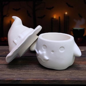 May include: A white ceramic Halloween-themed container with a witch hat lid. The container is shaped like a ghost with a star-shaped detail. The hat is off to the side, revealing the container's opening. The background is a dark, blurred scene.