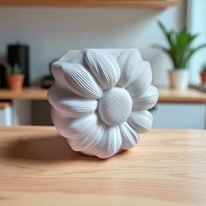 May include: A white, flower-shaped decorative vase. The vase has a detailed petal design and a textured center, resembling a sunflower. The vase is made of a solid material and sits on a wooden surface. Home decor.