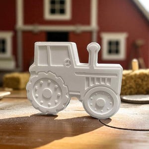 May include: A white tractor-shaped cookie cutter with detailed wheels, a cab, and a smokestack. The cutter is set on a wooden surface, with a red barn in the background. The image is well-lit, highlighting the cutter's design.