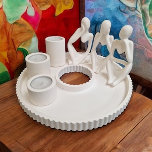 Könnte beinhalten: Weißer Kerzenhalter aus Keramik mit vier kleinen Kerzenbechern und drei abstrakten Figuren. Die Figuren sitzen um einen zentralen Kreis herum.
