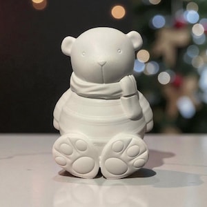 May include: A white ceramic bear figurine wearing a scarf and mittens. The bear is seated with its paws extended. The background is blurred with Christmas lights. The bear is a decorative item.