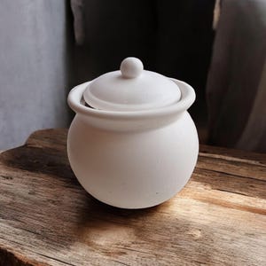 May include: A small, white ceramic pot with a lid, placed on a rustic wooden surface. The pot has a rounded shape with a small knob on the lid and two loop handles. The background is a soft, neutral colour.