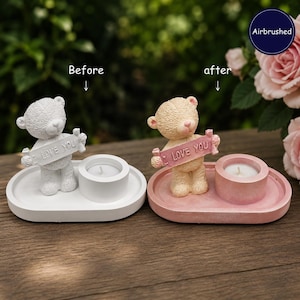 May include: Two oval candle holders with teddy bear figurines holding signs that read LOVE YOU. One holder is plain white, and the other is airbrushed in shades of pink. Each features a circular candle cup with a small white tea light candle inside.