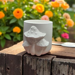 May include: A white ceramic planter with a raised mushroom design. The planter is cylindrical and sits on a weathered wooden surface. The background features colorful flowers in shades of yellow, orange, and pink.
