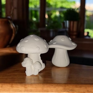 May include: Two white mushroom ornaments. One has a large cap with small, rounded bumps and a stem with three smaller mushrooms at the base. The other is a single mushroom with a smooth cap and stem. They are on a wooden surface.