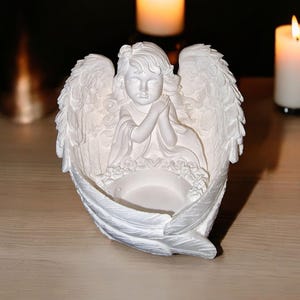 May include: White ceramic angel candle holder with a praying angel figurine in the center. The holder has a round opening for a tea light candle.