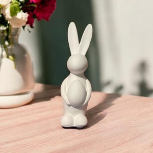 以下が含まれることがあります： 白い陶器製のウサギの置物で、白い卵を持っています。ウサギは薄い茶色の木製の表面の上に立っています。