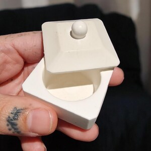 May include: A small white ceramic box with a lid. The lid has a small knob on top. The box is approximately 5 cm wide and 2.5 cm tall.