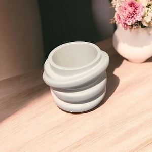 May include: A light blue ceramic planter with a unique, swirling design. The planter is made of smooth, textured ceramic and has a round shape.