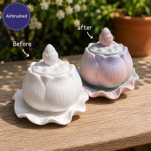 May include: Two ceramic lotus-shaped lidded jars on a wooden surface. The left jar is solid white. The right jar features an airbrushed gradient of soft pink, blue, and green. Both jars have a lotus bud finial on the lid and a scalloped base.