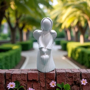 May include: A white ceramic angel figurine with outstretched wings and a heart-shaped object in its hands. The angel is standing on a brick wall, with a blurred background of trees and a pathway. Pink flowers are in the foreground.