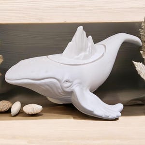 以下が含まれることがあります： 背中にスタイリッシュな山が乗った白いセラミック製のクジラの彫刻。クジラは空を泳いでいるように配置されています。