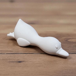 May include: A white ceramic duck figurine in a swimming pose. The duck has a long neck, a pointed beak, and small, round eyes. The figurine is lying on a wooden surface.