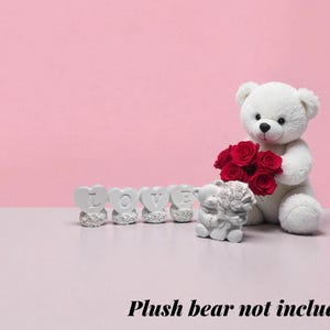 May include: A white teddy bear holding red roses, with heart-shaped ornaments spelling "LOVE". The background is pink. The text "Plush bear not included" is at the bottom of the image.