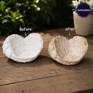 May include: Two angel wing bowls are displayed side-by-side. The left bowl is solid white, labeled "Before". The right bowl is white with gold-toned airbrushed highlights on the feather edges, labeled "after". Both bowls feature a curved, heart-like shape.