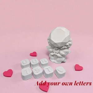 May include: A white figurine of an angel holding a teddy bear, with the words "OUR 1ST" spelled out in white letter blocks. Several pink heart-shaped decorations are scattered around the objects on a pink background. The text "Add your own letters" is at the bottom.