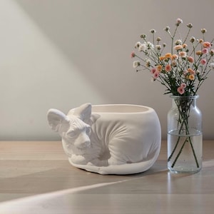 May include: A white ceramic cat-shaped planter and a clear glass vase with flowers. The planter has a detailed cat design. The vase contains a bouquet of small, colorful flowers. The objects are on a wooden surface, creating a simple, elegant display.