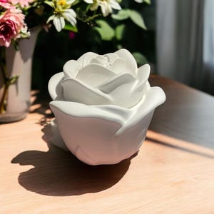 Peut inclure: Objet décoratif en forme de rose en céramique blanche. La rose est posée sur une surface en bois.