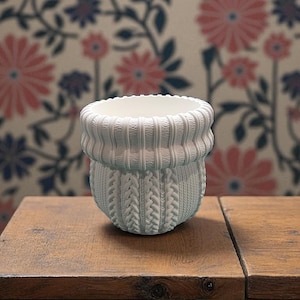 May include: A white ceramic planter with a knitted sweater design. The planter has a ribbed top and a cable-knit pattern on the body. It sits on a wooden surface against a floral wallpaper background.
