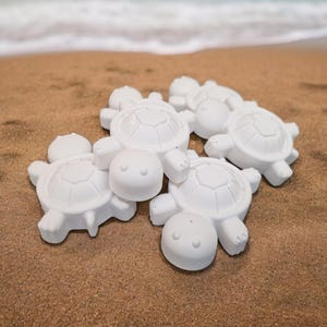 May include: Several white turtle figurines are arranged on sand, with the sea and shoreline in the background. The turtles feature detailed shell patterns, small heads, and limbs. The scene evokes a beach theme.