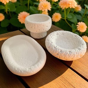 May include: Three white, textured, ceramic pieces sit on a wooden surface. The pieces include an oval dish, a pedestal bowl, and a round bowl. Peach-colored flowers and green foliage are in the background.