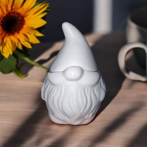 May include: A white ceramic gnome figurine with a pointed hat and a beard. The gnome is sitting on a wooden surface.