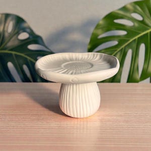May include: A white ceramic mushroom-shaped decorative piece. The mushroom has a rounded cap with a textured surface and a ribbed stem. The piece is set on a light-colored wooden surface, with green leaves in the background.