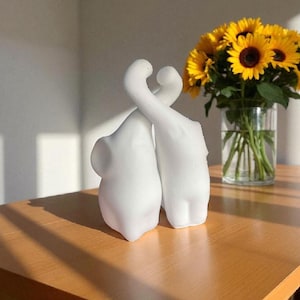 以下が含まれることがあります： 木製のテーブルの上に置かれた、絡み合った形の白い抽象彫刻のペア。背景には、黄色の花びらと濃い茶色の中心を持つヒマワリの花瓶があり、シーンに彩りを添えています。