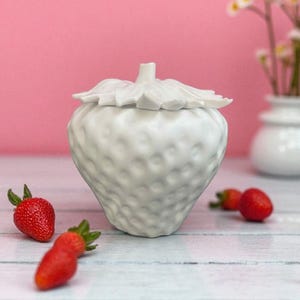 以下が含まれることがあります： ピンク色の背景に、白いイチゴ型のセラミック容器と蓋が置かれています。新鮮なイチゴが容器の周りに散らばっています。背景には、花が入った小さな白い花瓶があります。