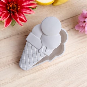 May include: A white silicone ice cream cone mold with two scoops of ice cream. The mold is on a light wood surface.