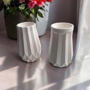 以下が含まれることがあります： 幾何学模様の白とグレーの装飾用花瓶2つ。花瓶は円筒形で、下部は多面カットされています。背景にはフラワーアレンジメントがあります。