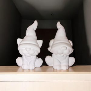 May include: Two white ceramic cat figurines wearing pointy hats. The cats are sitting on a white shelf.