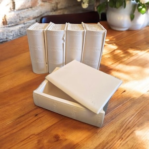 May include: A white ceramic box with a lid that looks like a book. The box is sitting on a wooden table with a stack of four books behind it.