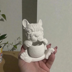 May include: A white ceramic bulldog figurine, holding a round object. The bulldog is sitting with its paws on the object. The figurine has detailed facial features and is unpainted.