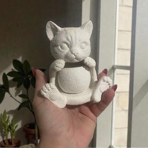 May include: A white ceramic cat ornament with a round belly, paws, and a head with pointed ears. The cat is holding its belly with its paws. The ornament is unpainted and appears to be a planter or decorative item.