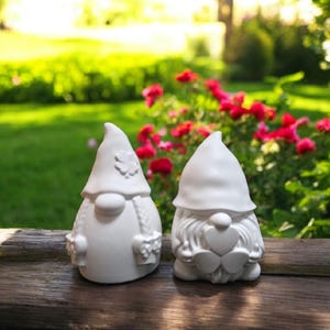 May include: Two white ceramic garden gnomes with pointed hats and detailed features. One gnome has a flower on its hat and baskets, the other has a heart-shaped beard. They are set on a wooden surface with a blurred garden background.