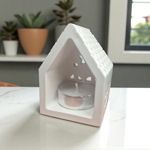 May include: A white ceramic house-shaped candle holder with a textured roof and star-shaped cutouts. The candle holder has a pink ombre effect at the base and holds a lit tea light candle. The background includes plants and a window.