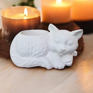 May include: White ceramic cat candle holder featuring a sleeping cat design. The cat has its eyes closed and is curled up in a sleeping position. The holder has a small opening at the top for a tea light candle.