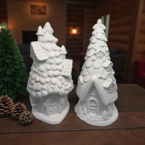 May include: Two unpainted ceramic Christmas village houses with a tree-like design. The houses have detailed features like doors, windows, and textured roofs. A small green Christmas tree and pine cones are also on the wooden table.