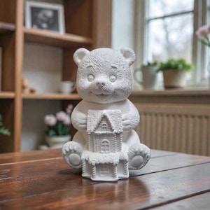 May include: A white ceramic bear ornament holding a detailed house. The bear has a smiling face and is sitting on a wooden table. The background includes a bookshelf, window, and plants.
