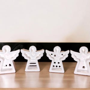 Könnte beinhalten: Vier weiße Keramik-Engelfiguren mit verschiedenen Mustern auf ihren Kleidern. Die Engel stehen auf einer Holzoberfläche.