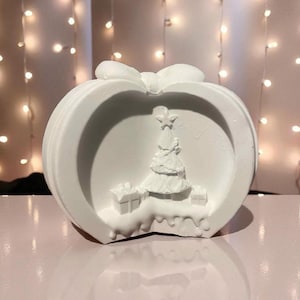 Peut inclure: Ornement de Noël blanc et rond avec un nœud sur le dessus. L'ornement présente un relief détaillé d'un sapin de Noël et de cadeaux à l'intérieur d'un cadre circulaire. L'ornement est posé sur une surface rose.