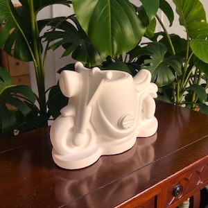 May include: A white ceramic motorcycle planter with a headlight, wheels, and seat. The decorative planter is placed on a dark brown wooden table. Green leafy plants are in the background.