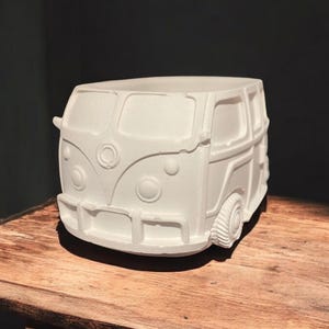 May include: A matte white planter shaped like a vintage camper van. The vessel features molded details including a front bumper, circular headlights, a central emblem, and side windows. The top is open, providing a hollow interior space.