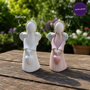 May include: Two angel figurines are displayed side by side. The left figurine is solid white. The right figurine is airbrushed with soft pink and blue pastel tones. Both figures feature a smooth oval face, wings, and hold a heart-shaped object.