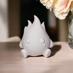 May include: A small, white, cartoon-like figurine shaped like a flame. It has a smiling face, small arms and legs, and is sitting on a light-colored wooden surface. The background includes a vase of flowers.