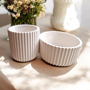 May include: Two white ceramic planters with a ribbed design. The larger planter is wider and shallower than the smaller planter.