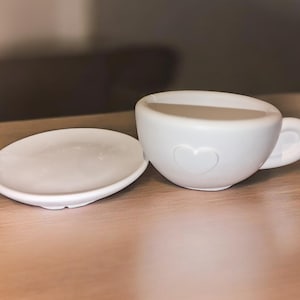 May include: A white ceramic teacup and saucer set with a heart design. The cup has a handle and a wide rim. The saucer is round and has a slightly raised edge.